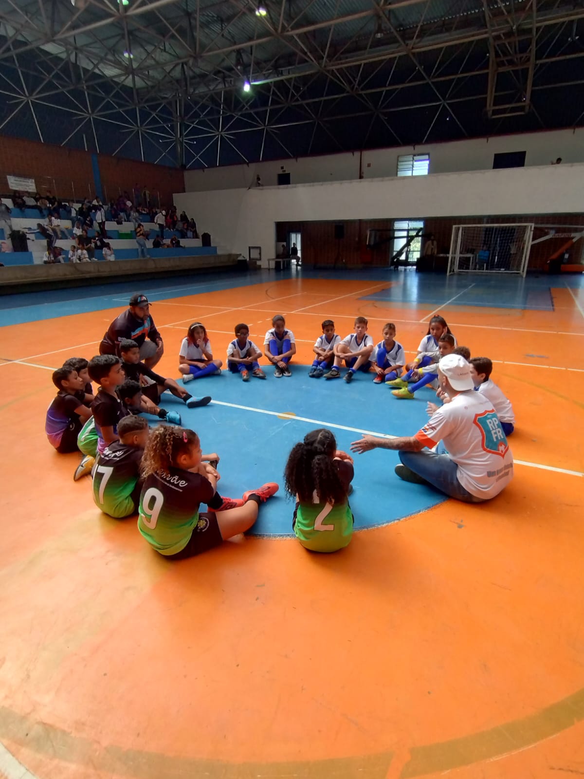 Na imagem, os dois times estão reunidos em círculo na quadra, sentados com os treinadores, conversando sobre o jogo que realizaram